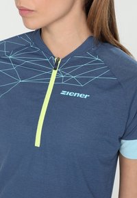 Modré sportovní tričko s krátkým rukávem a žlutým zipem. Má geometrický vzor světle modré barvy na rameni a logo na hrudi.