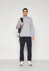 Világosszürke hosszú ujjú ing cipzáros gallérral, fekete nadrág és fehér tornacipő. Egy fekete hátizsák piros részletekkel az egyik vállon van hordva.