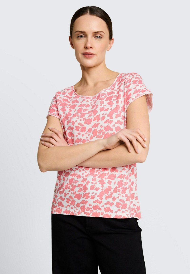 Tumedate juustega naine roosa-valge lillelise lühikeste varrukatega särgi ja mustade pükstega, seisab käed rüpes ühevärvilise tausta ees.