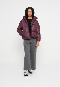 Vrouw die staat in een paarse pufferjack, zwarte top, grijze wijde cargobroek, witte sokken en zwarte sneakers voor een witte achtergrond.