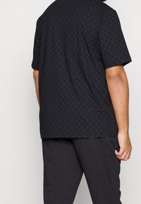 Camisa negra de manga corta con un patrón de logotipos repetido, que presenta un corte relajado y una textura suave, combinada con pantalones oscuros.