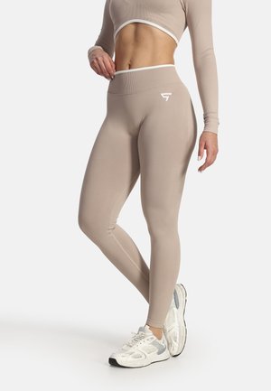 Frau trägt beigefarbene High-Waist-Leggings und langärmliges Crop-Top, richtet den Bund, steht in weißen Sportschuhen vor weißem Hintergrund.