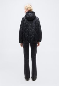 Άτομο με κοντά ξανθά μαλλιά που φορά μαύρο μπουφάν με κουκούλα και φερμουάρ και σκούρα γκρι παντελόνια με φουντάκια, στέκεται με την πλάτη γυρισμένη σε λευκό φόντο.