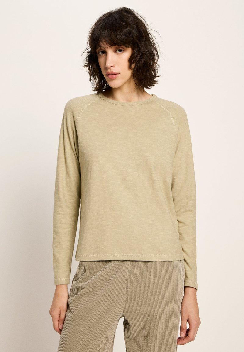 Frau mit kurzen, welligen Haaren, die ein beiges Langarmoberteil und eine hellbraune Cordhose trägt, steht vor einem schlichten, hellen Hintergrund.