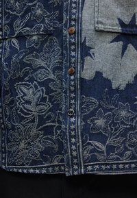 Τζιν πουκάμισο με ανθικό και αστέρια σχέδια, διαθέτει καφέ και μαύρα κουμπιά κατά μήκος της κεντρικής ραφής και μια τσέπη στη δεξιά πλευρά.