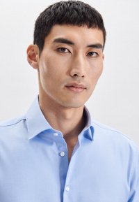 Jasnoniebieska koszula z długim rękawem o teksturowanej tkaninie, z spiczastym kołnierzykiem i białymi guzikami. Posiada subtelny kontrastujący wewnętrzny kołnierzyk.