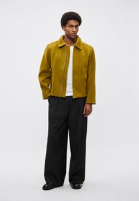 Volnena suknja gorčične barve z zadrgo in strukturiranim ovratnikom, v kombinaciji z ohlapnimi črnimi hlačami in črnimi čevlji.