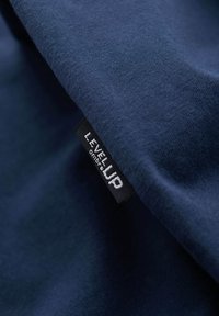 Gros plan sur un tissu bleu foncé avec une étiquette de vêtement noire cousue dans la couture, portant l'inscription "LEVEL UP" en lettres blanches.