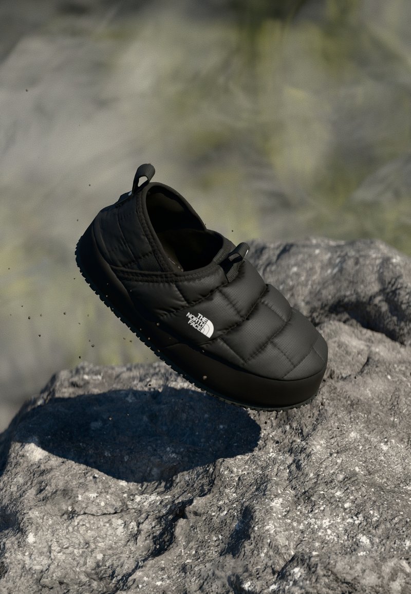 Scarpa slip-on nera trapuntata con suola testurizzata, caratterizzata da una punta arrotondata e un logo del marchio sul lato, posizionata su uno sfondo roccioso.