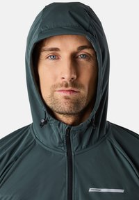 Giacca a vento impermeabile di colore verde scuro con zip frontale, cordino regolabile sul cappuccio e logo bianco sul lato sinistro.