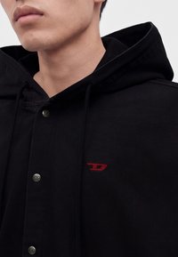 Gros plan d'une personne portant une veste à capuche noire avec des boutons-pression en métal et un petit logo rouge brodé sur la poitrine.