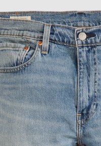 Gros plan sur un jean en denim bleu montrant les poches avant, les passants de ceinture, le bouton en métal et les rivets en cuivre sur un fond clair.