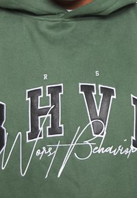 Zielona bluza z kapturem z wyraźnymi, czarnymi literami "BHV" z białym przeszyciem. Poniżej znajduje się napis "Worst Behavior" w stylu odręcznym.