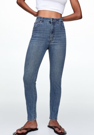 Jean bleu slim taille haute porté avec des tongs noires et un débardeur court blanc, debout les mains sur les hanches.