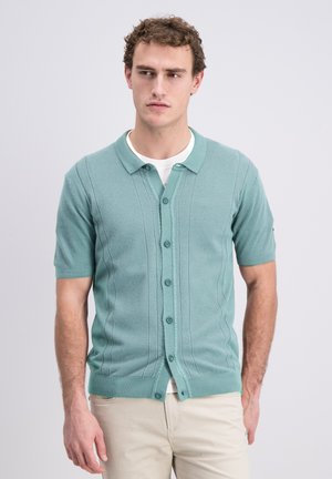 Jeune homme en cardigan vert menthe à manches courtes boutonné sur une chemise blanche et un pantalon beige, debout devant un fond uni.