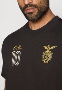 Camiseta de algodón negra con mangas cortas. Presenta el texto bordado en amarillo "O Rei" y el número "10"; incluye un emblema con diseño de águila y escudo.
