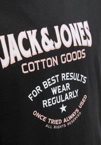 Juodas medvilnės marškinėlis su dideliais baltais ir rožiniais tekstais. Tekste yra "JACK & JONES", produkto detalės ir žvaigždutė.