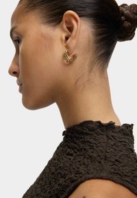 Profiel van een vrouw met een gevlochten haarbun, die een gestructureerde donkere top en een gouden oorring met verhoogde ontwerpdetails draagt.
