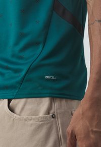 Tealfärgad träningsskjorta med en texturerad tygstruktur och ett upprepande mönster samt en svart accentrand. "DRYCELL"-etikett synlig på sidan.