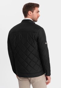 Sort quiltet jakke med en glat tekstur, der har et subtilt diagonalt mønster og et lille logo på venstre ærme.