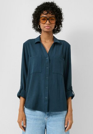 Vrouw met krullend haar draagt grote amberkleurige zonnebril, donker petrolgroene blouse met opgerolde mouwen en lichtblauwe spijkerbroek, staand tegen een witte muur.