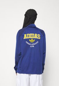 Mörkblå långärmad jacka med krage, med gul och vit text på baksidan: "ADIDAS ATHLETIC CLUB" och Adidas-logotypen.