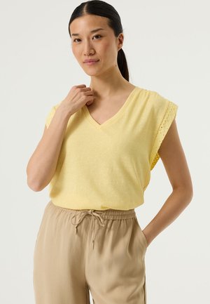 Camiseta amarilla de cuello en V con detalles de encaje texturizado en las mangas, combinada con pantalones beige con cordón, destacando tela suave y ajuste relajado.