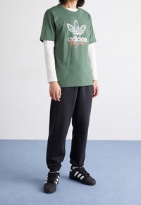 Zelené bavlněné tričko s bílým logem Adidas a červenými akcenty, přes bílé tričko s dlouhým rukávem. V kombinaci s černými tepláky a teniskami.