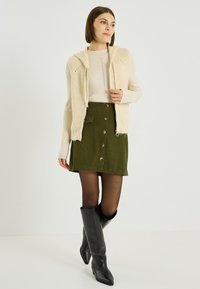 Gilet en maille crème, haut à manches longues côtelées, jupe en velours côtelé vert olive avec boutons à l'avant, et bottes en cuir noires jusqu'aux genoux.