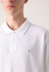 Hvit polo skjorte med klassisk krage, med to knapper og en liten grå "CK"-logo på venstre bryst. Teksturt stoff.