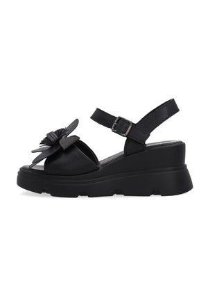 Czarna sandałka na koturnie z grubą podeszwą, regulowanym paskiem wokół kostki i ozdobą w postaci kwiata na przednim pasku, widziana z boku.