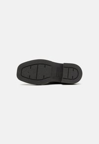 Suela de zapato de goma negra con patrón de agarre texturizado. Presenta una forma redondeada y un logo grabado. Acabado suave con surcos definidos.