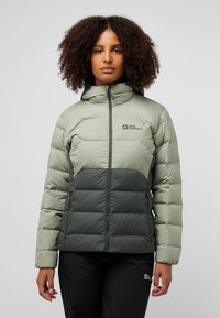 Giacca a vento verde e verde scuro con chiusura a zip, caratterizzata da un design imbottito e un logo discreto sulla parte superiore sinistra del petto.