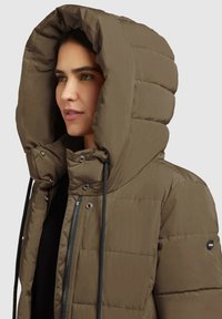 Brūns pūka jaka ar lielu, quiltētu kapuci. Ar rāvējslēdzēju priekšā, spiedpogām un auklām. Materiāls ir gluds ar elegantu apdari.