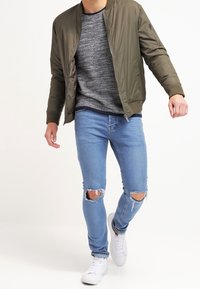 Bomber bunda olivově zelené barvy přes šedý pletený svetr, doplněná světle modrými roztrhanými džínami a bílými teniskami. Neformální venkovní outfit.