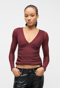 Γυναίκα με πλεγμένα μαλλιά, που φοράει εφαρμοστή μακρυμάνικη μπορντό μπλούζα με V-λαιμόκοψη και μαύρο δερμάτινο παντελόνι, με τα χέρια στις τσέπες, στέκεται μπροστά σε λευκό φόντο.