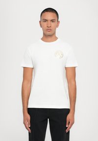 Hvit bomull T-skjorte med rund hals og korte ermer, med et sirkulært gulllogo på venstre bryst. Enkelt og klassisk design.