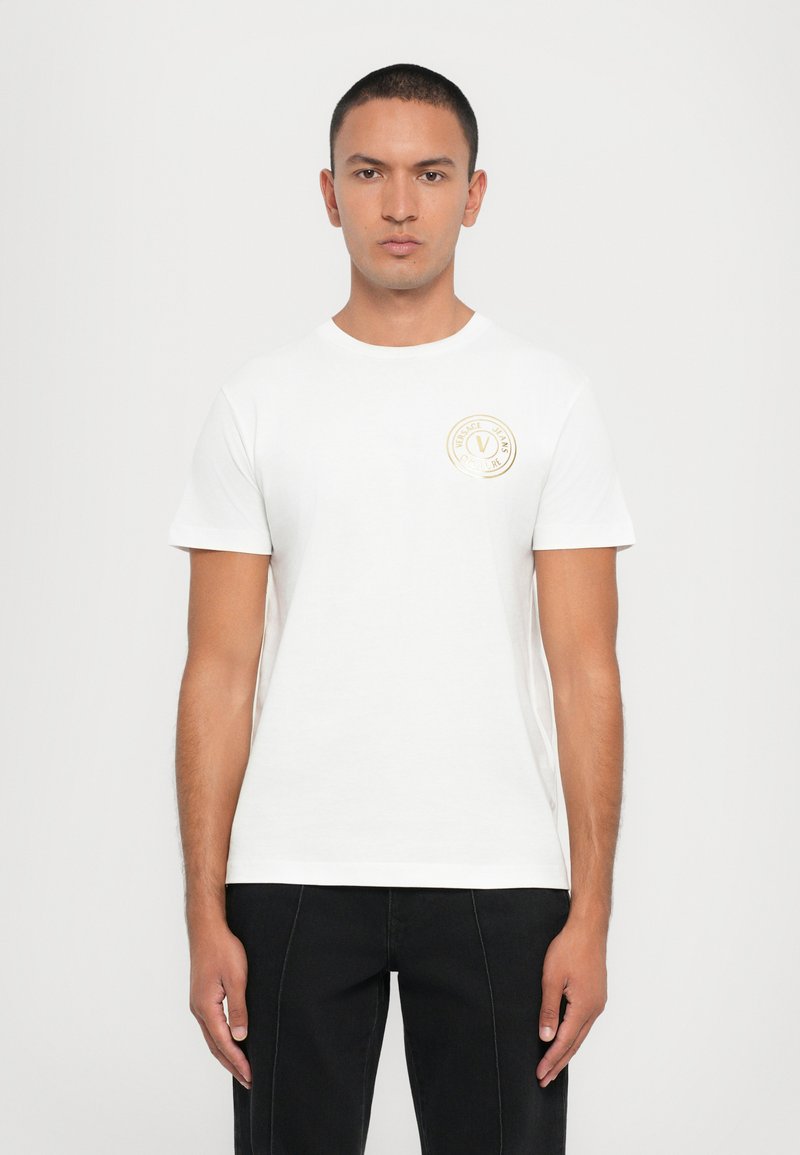 Hvit bomull T-skjorte med rund hals og korte ermer, med et sirkulært gulllogo på venstre bryst. Enkelt og klassisk design.