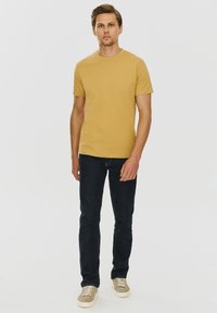 Musztardowa żółta koszulka z krótkim rękawem, w połączeniu z ciemnoniebieskimi dżinsami i beżowymi trampkami, stojąca na białym tle.