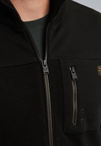 Mustat vetoketjulliset takki tekstuuripinnalla, näkyvät vetoketjut ja rintatasku. Ominaisuudet kultaisista yksityiskohdista ja korkea kaulus.