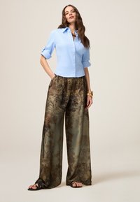 Helles blaues Hemd mit hochgerollten Ärmeln, kombiniert mit weit geschnittenen, gemusterten braunen und grünen Hosen. Schwarze Sandalen und ein Armband.