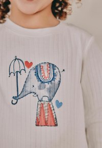 Biała, prążkowana koszulka z kolorowym haftowanym wzorem przedstawiającym słonia balansującego na czerwono-niebieskim przedmiocie, trzymającego parasolkę.