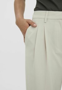 Pantalons plissés beige clair avec une coupe ajustée. Présentent des poches latérales et une texture de tissu lisse, mis en valeur à côté d'une main reposant sur la poche.