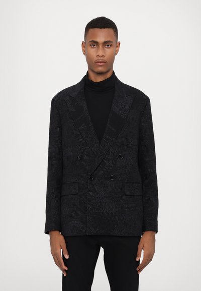 Double-breasted black blazer with textured woven pattern, lapel collar, two front pockets, and tailored fit. Worn over a black turtleneck.