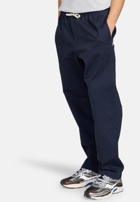 Pantalon en coton bleu marine avec taille élastique et cordon de serrage. Caractéristiques des poches latérales et une coupe décontractée. Associé à des baskets argentées et noires.