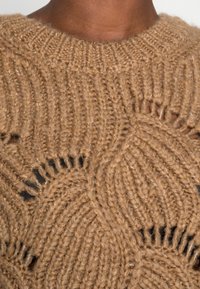 Pull en tricot couleur fauve avec un motif texturé comprenant des sections circulaires ouvertes. Encolure côtelée et matière douce et épaisse.