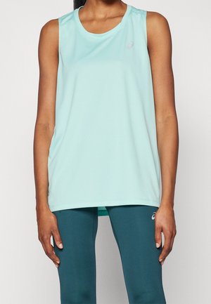 Frau trägt ärmelloses helltürkisfarbenes Sporttop und dunkelgrüne Leggings und steht vor einem schlichten hellen Hintergrund.