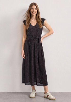 Femme souriante portant une robe midi noire sans manches avec des épaules à volants et des baskets blanches, debout devant un mur clair uni.