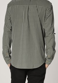 Tył osoby ubranej w zielono-czarną koszulę w kratę z długim rękawem i czarne spodnie, ręce w kieszeniach.