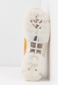 Semelle de sneaker en caoutchouc blanc translucide avec des formes étoile et croix métalliques intégrées, montrant le motif de la bande de roulement et le bord de la chaussure en jaune et rose.